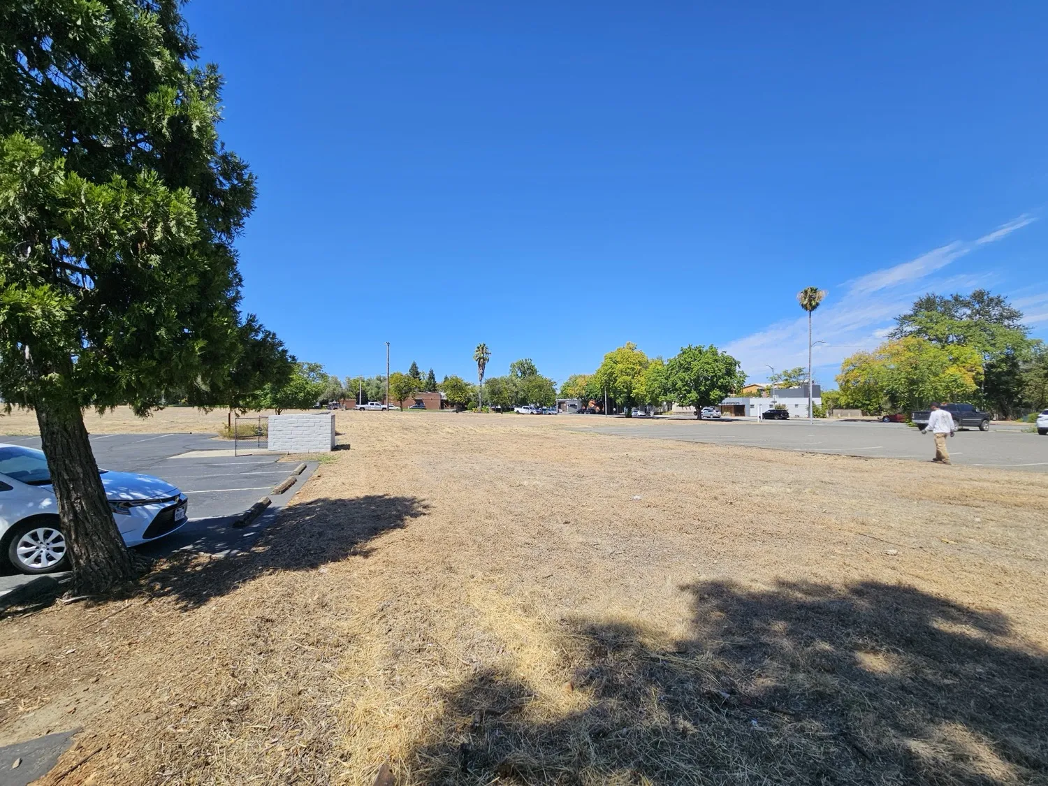 Corner lot cut down to short dry grass with a car parked at the edge and a parking lot beyond