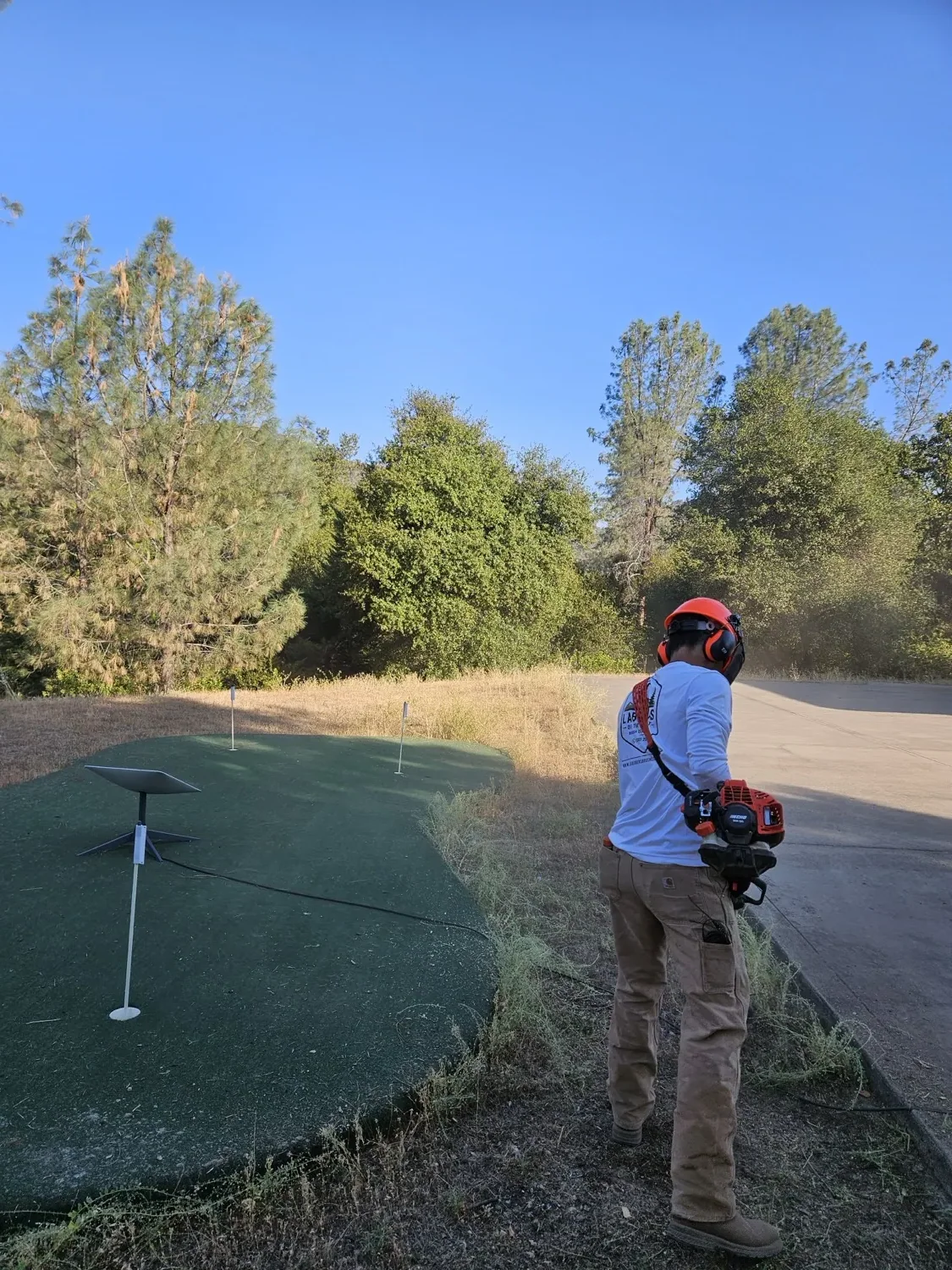 Crew member in safety gear cutting overgrowth beside a backyard putting green